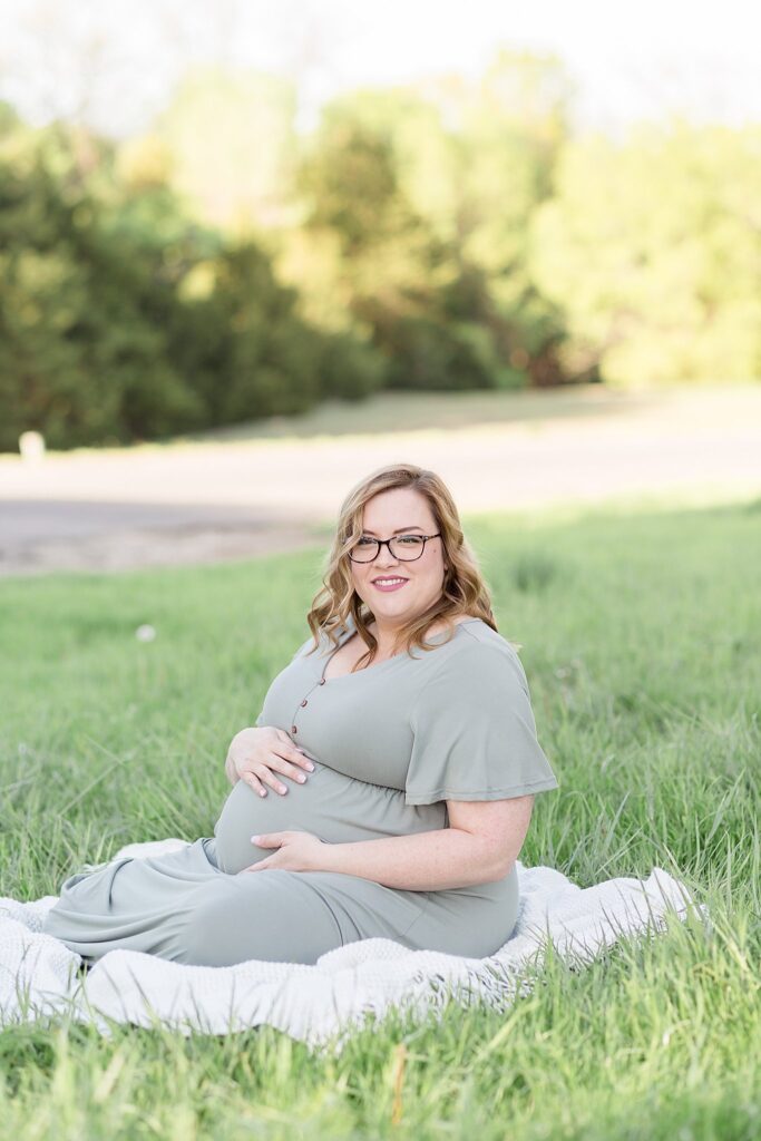 Pregnant mom sits on white blanket on the grass at Erwin Park in McKinney, TX as she holds her belly and looks off into the distance.