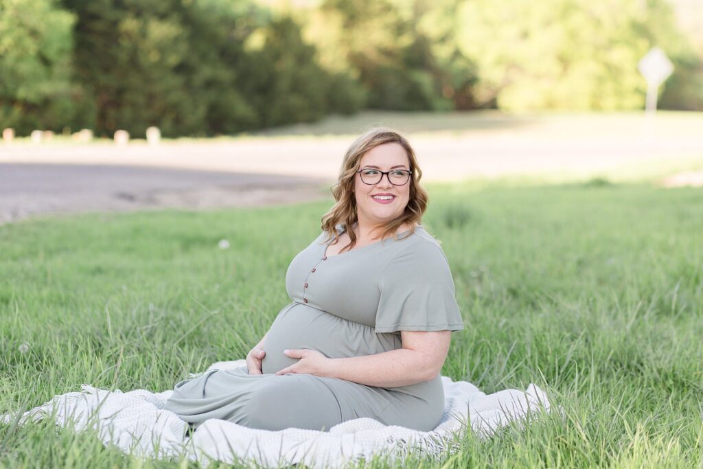 Pregnant mom wears a muted mint/grey long dress as she holds belly and sits on a white blanket in the grass.