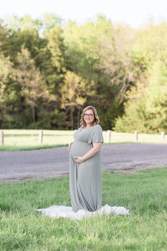 Wisp + Willow Photography Co. has a maternity portrait session in McKinney, TX at Erwin Park on a beautiful day with the sun hitting the tress in the background as pregnant mom stands in the shade on the grass and looks at camera.