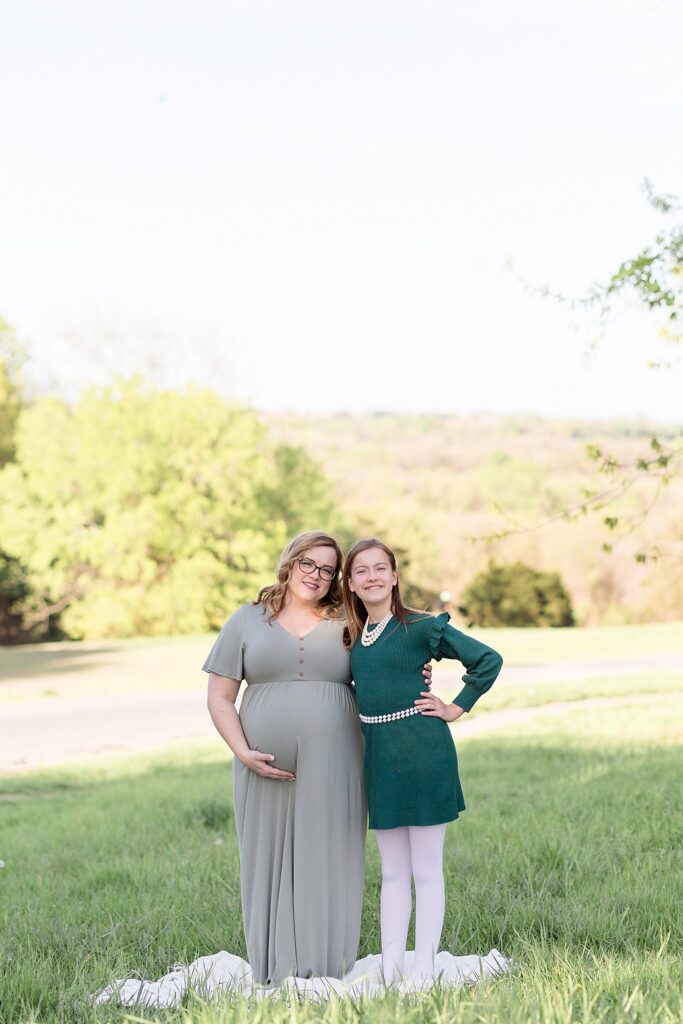 Pregnant mom stands with young daughter in their coordinating green colored dresses as they smile at the camera of Wisp + Willow Photography Co. during a maternity portrait sesion.