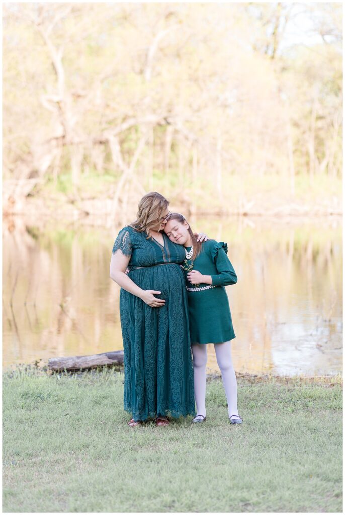 Pregnant Mom stands wearing an emerald green lacey dress as she hugs daughter who rests her head on her mom's shoulder during their photography session in McKinney, TX.