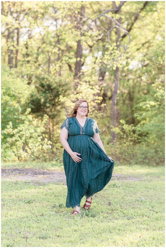 Wisp + Willow Photography Co. have a maternity portrait session at Erwin Park in McKinney, TX as pregnant mom walks in the grass and smiles off in the distance as she holds her belly with one hand and her dress out in the other.