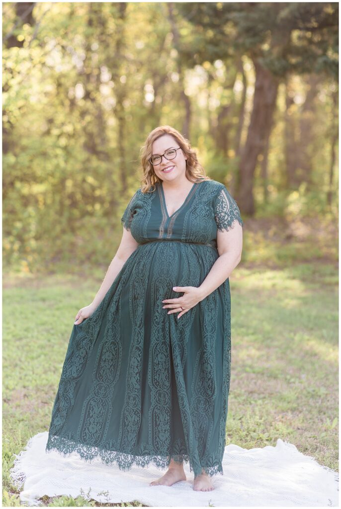 Pregnant mom wears beautiful green, lace, long dress for her maternity portrait session in McKinney, TX.  She smiles at the camera while holding her belly with one hand and her dress with the other.