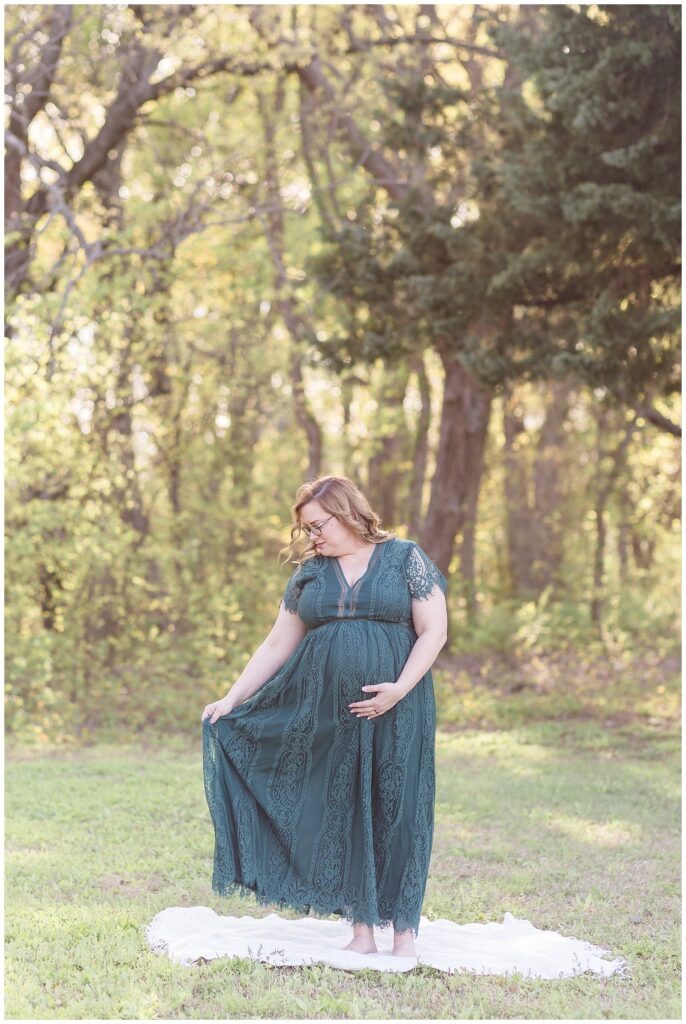 Maternity portrait session in McKinney, TX captures beautiful mom-to-be wearing an emerald, v-neck, lace, long dress while mom holds belly and looks down at her dress she is holding with the other hand.