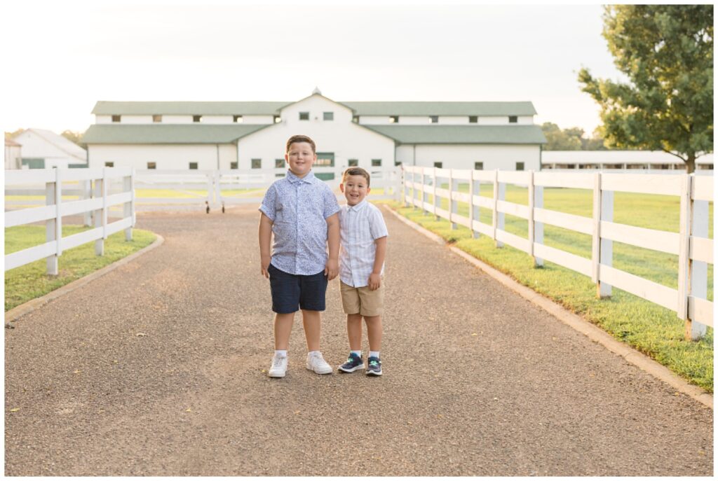 Two brothers wear button up shirts and shorts with tennis shoes as they smile at the camera standing in front of a barn.