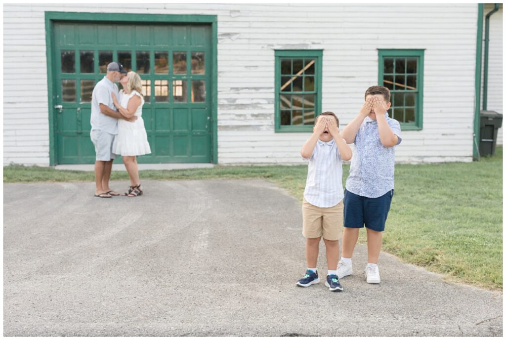 Two sons cover their eyes with their hands as they stand in the forefront of their parents who sneak a kiss behind them during their family session with Wisp + Willow Photography Co.