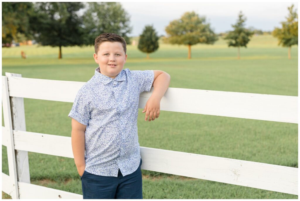 Young boy rests his arm on white fence with his other hand in his pocket as he smiles at the camera wearing blue and white during his Franklin family session.