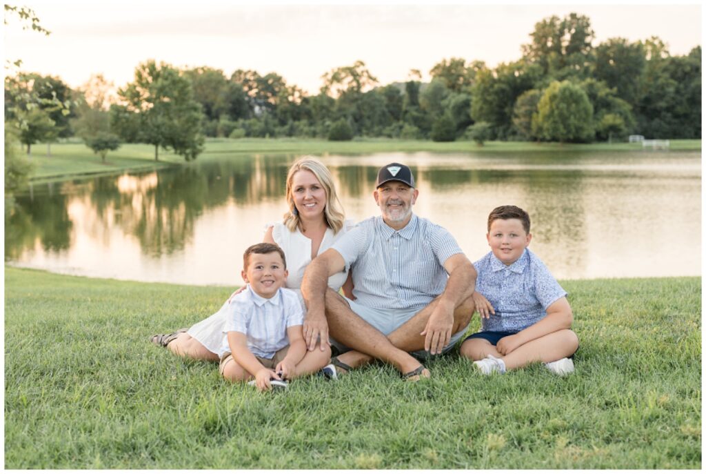 Family of 4 sit in grass in front of a pond during sunset as they smile at the camera of Wisp + Willow Photography Co. during their Franklin family session at Harlinsdale Farm in TN.