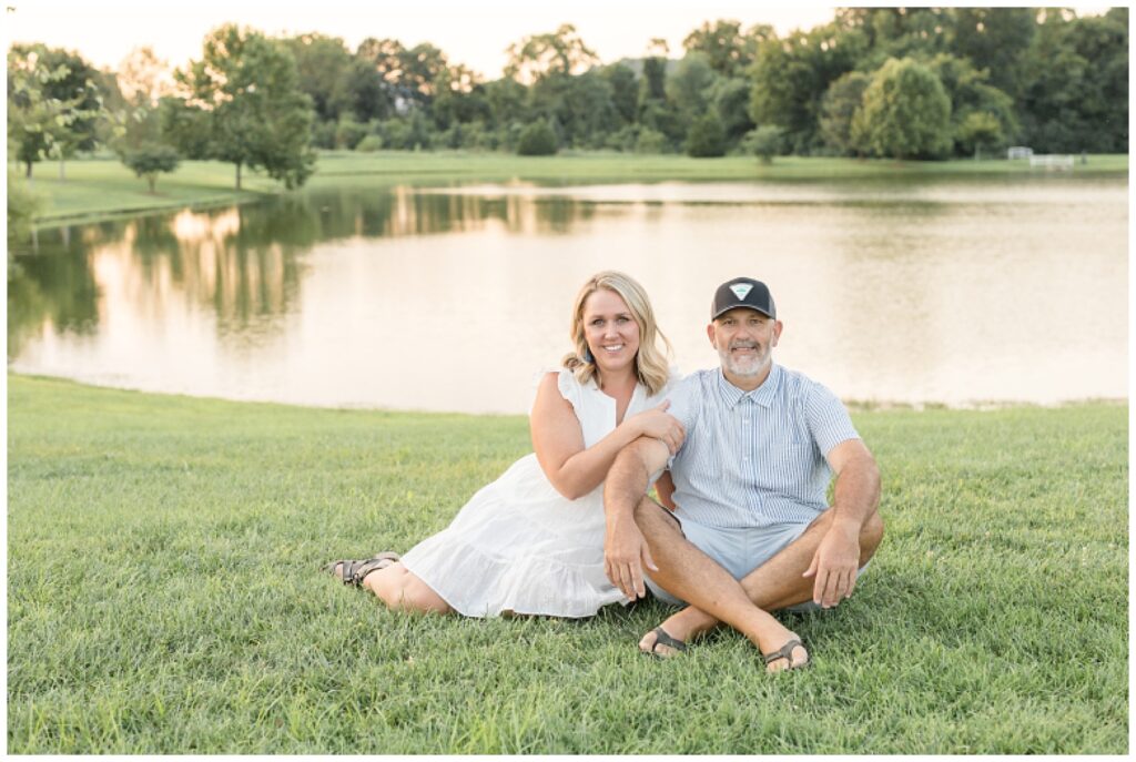 Husband and wife sit in front of pond on the grass in Franklin, TN as the white holds on to husbands arm and they smile at the camera.