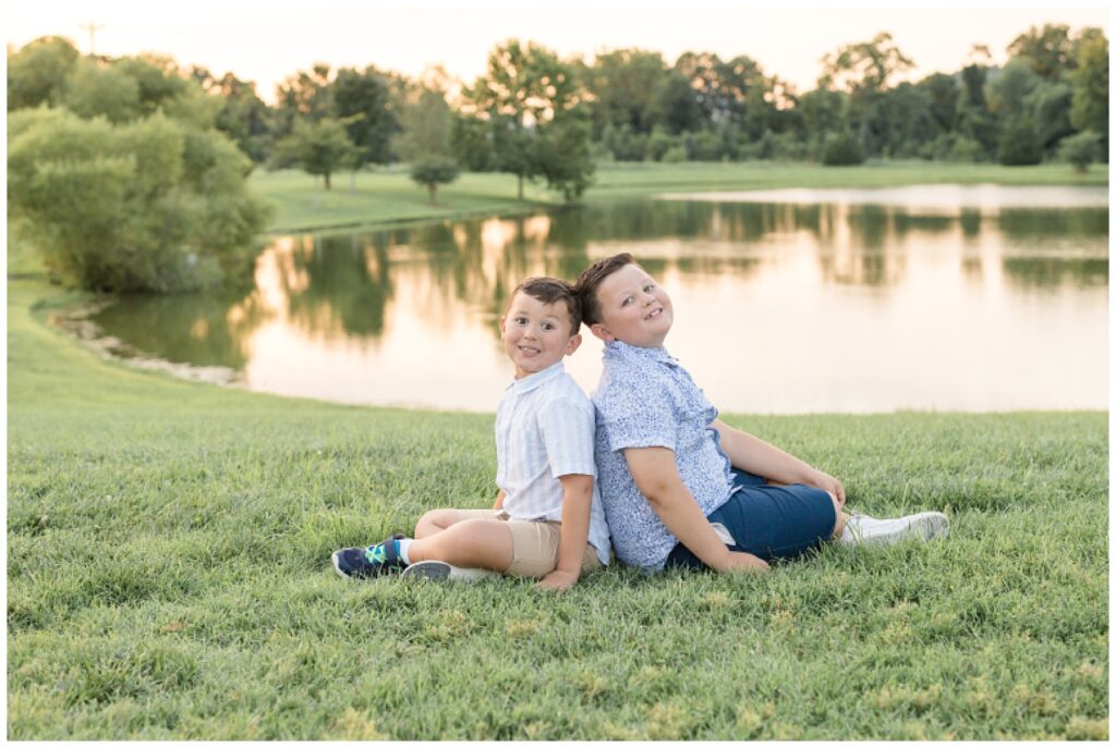 Two sons sit back to back during their Franklin family session at Harlinsdale Farm wearing coordinating blue and white outfits as they sit in the grass in front of a pond at sunset.