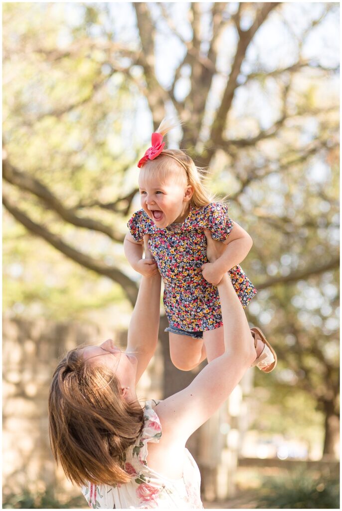 Mom holds up toddler girl who wears a pink bow headband and a floral shirt and jean shorts during their family session in Frisco, TX.