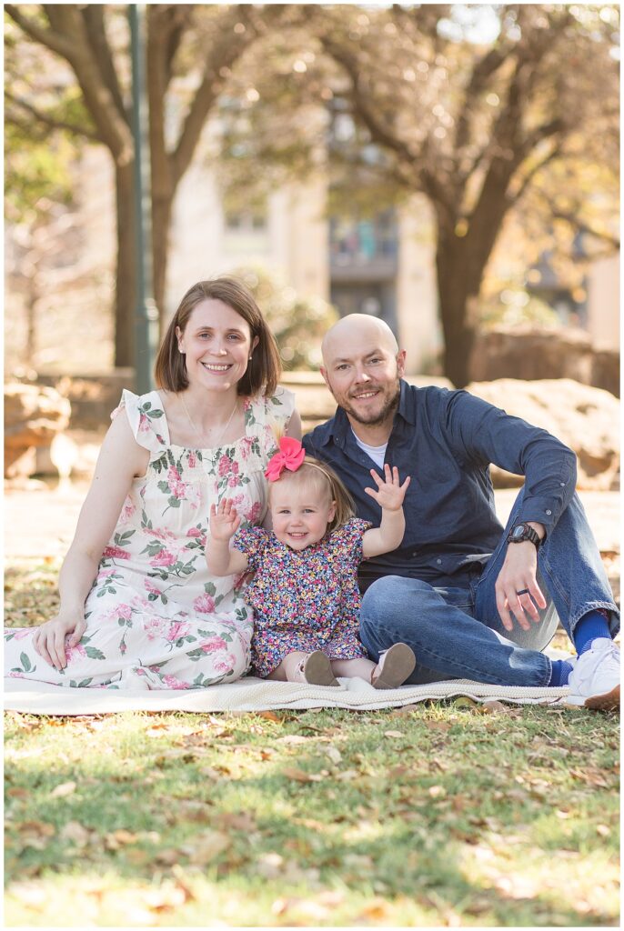 Mom and Dad sit in Frisco Central park with their toddler daughter who sits between them as she puts her hands up in the air and they all smile at the camera.