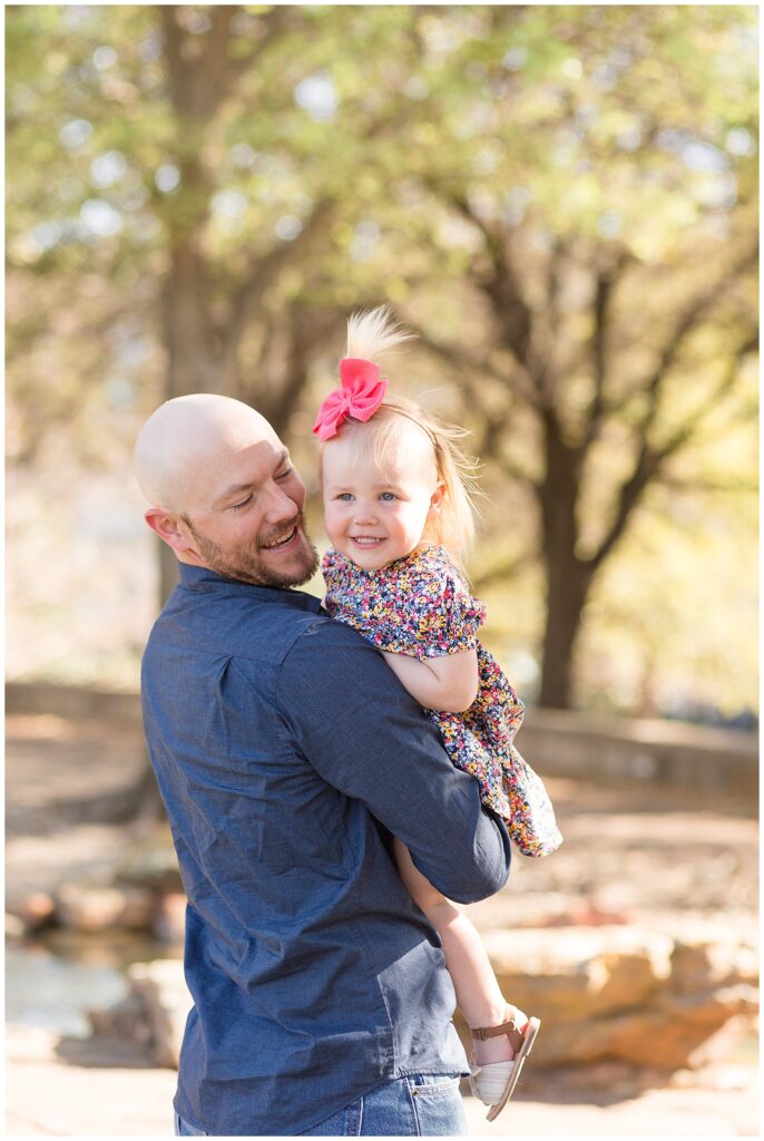 Dad holds daughter as he looks at her and daughter looks off past the camera in her pink bow headband and floral shirt during their family session with Wisp + Willow Photography Co.