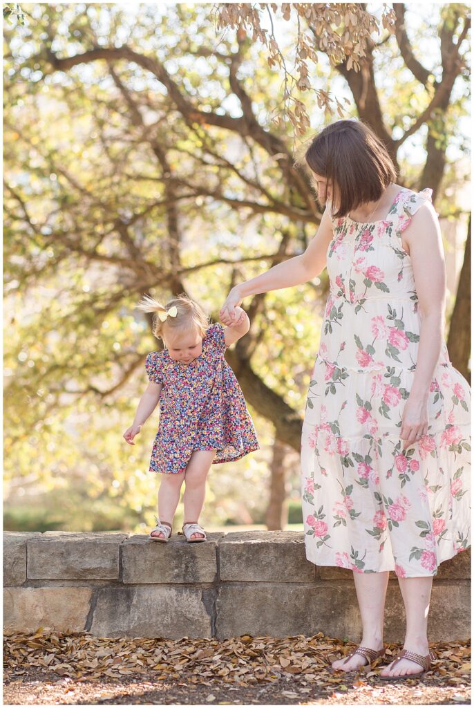 Mom holds hand of toddler daughter as she walks along a rock wall and looks to jump.  Both mom and daughter wear floral patterned dresses for their family session in Frisco, TX.