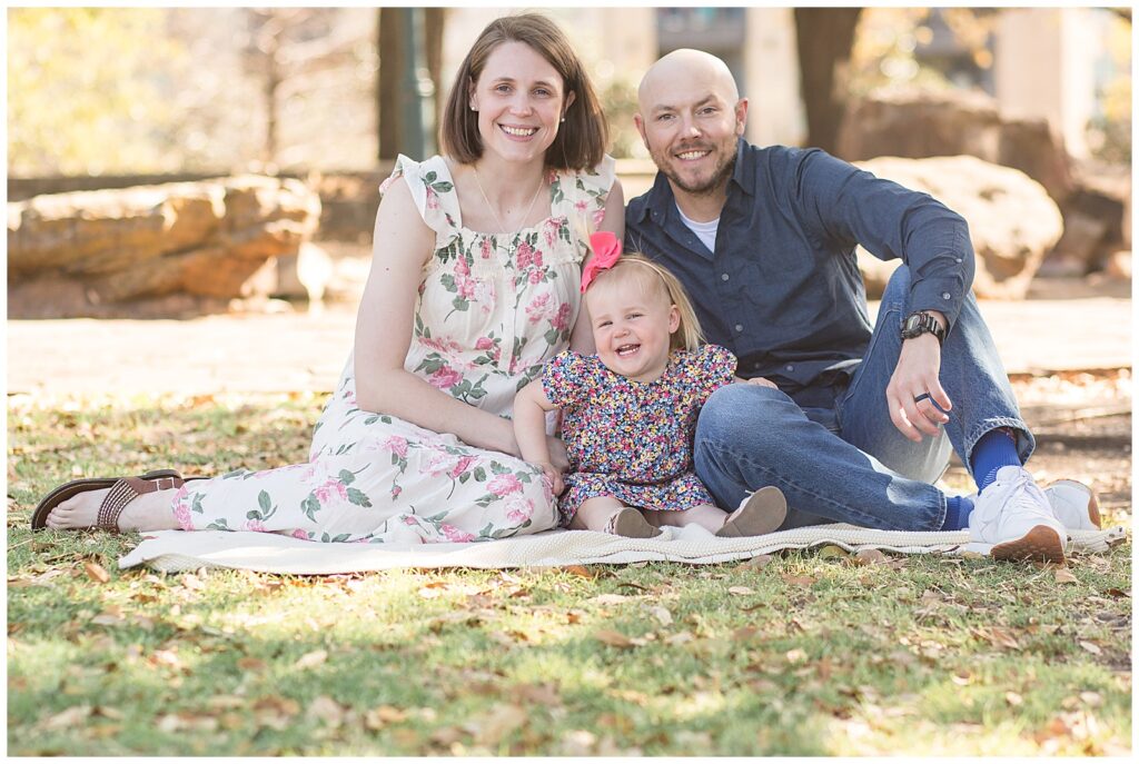 Family of 3 sit on the ground of Frisco Central Park as they smile at the camera of Frisco family photographer, Wisp + Willow Photography Co. during their family session.