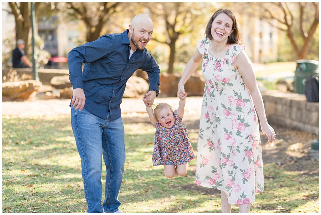 Mom and Dad swing toddler girl in the park of Frisco Central for a fun candid moment captured by Wisp + Willow Photography Co.