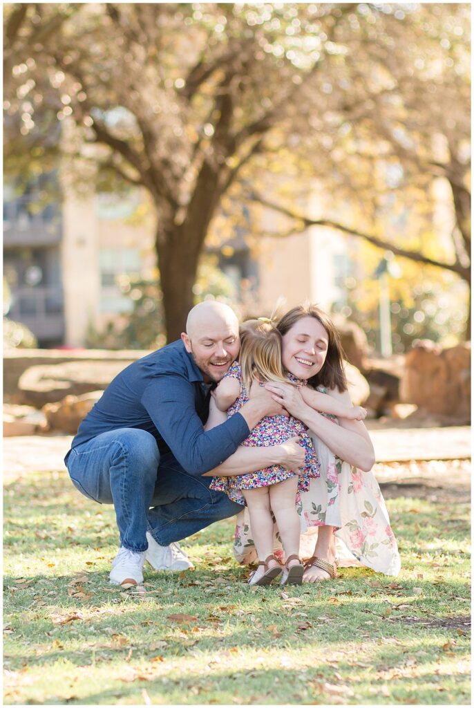 Toddler girls runs to Mom and Dad and gives them the biggest hug at Frisco Central Park during their family portrait session.  They wear coordinating outfits of floral and Dad wears a solid blue button down with jeans.
