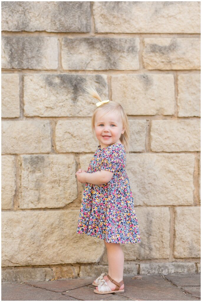 Toddler girl with blonde hair and blue eyes stands in front of a rock wall and smiles at the camera with her floral dress and yellow bow.