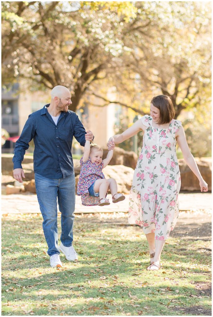 Mom and Dad hold hands of toddler girl in the park of Frisco Central Park as they swing her in front of Frisco family photographer, Wisp + Willow Photography Co.  Check out the blog to see more!