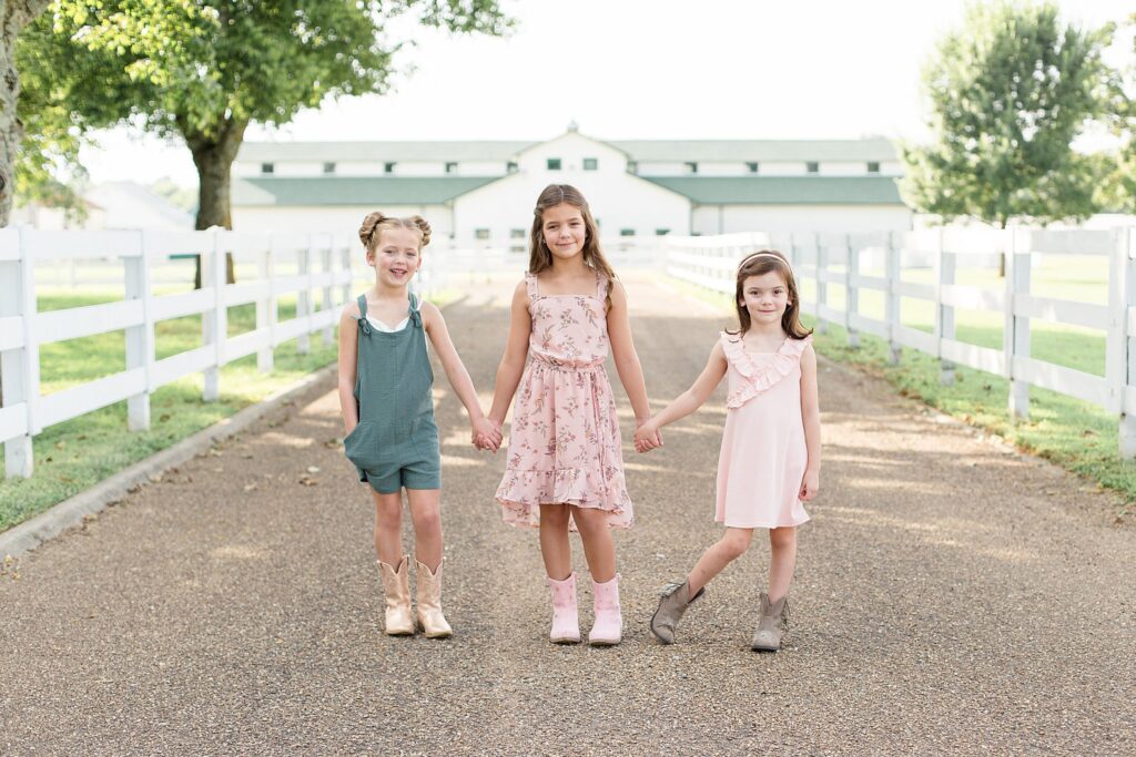 3 sisters stand on the driveway of Harlinsdale Farm in Franklin, TN wearing coordinating outfits of pink and teal and cowgirl boots!  They are holding hands and looking at the camera of Wisp + Willow Photography Co.