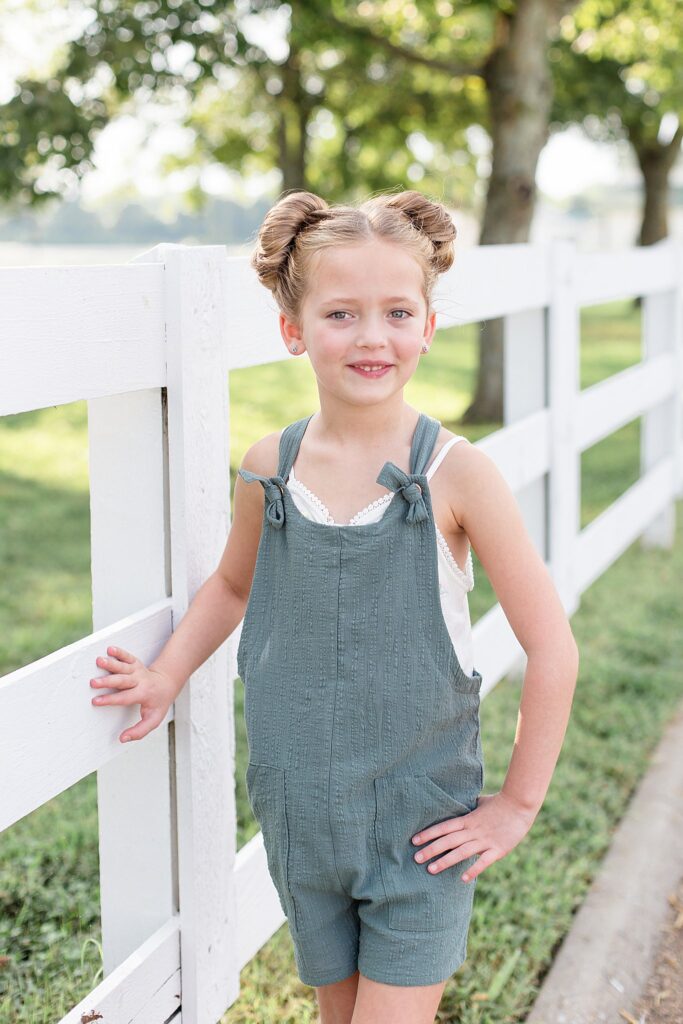 Little girl wears two buns, a teal-ish romper, and a white spaghetti strap tank underneath, as she smiles at the camera of Wisp + Willow Photography Co.,with one hand on a white fence and the other hand on her hip.