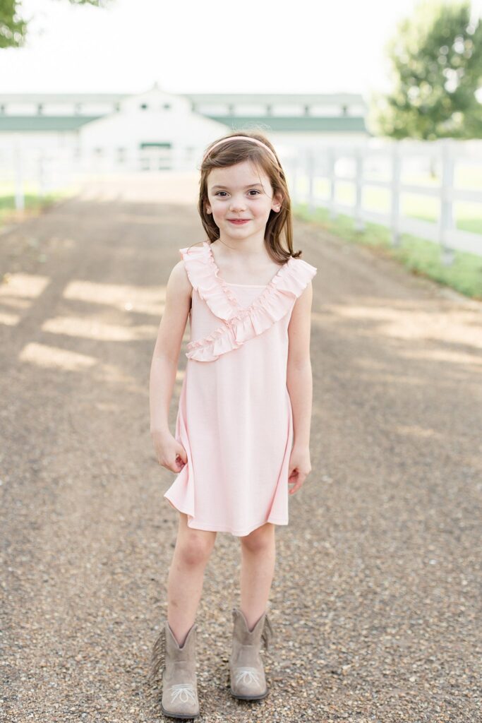 Little girl stands on the driveway of Harlinsdale Farm in Franklin, TN, wearing a light pink dress and a light pink thin headband as she smiles at camera during her sibling photography session.