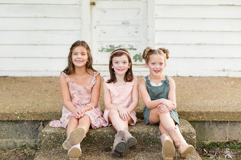 2 sisters where light pink dresses, one solid and the other floral, while the third sister wears a teal-ish romper and they all wear cowgirl boots.  They are sitting on the stoop of an old farmhouse with their legs crossed and smiling at the camera of Wisp + Willow Photography Co.