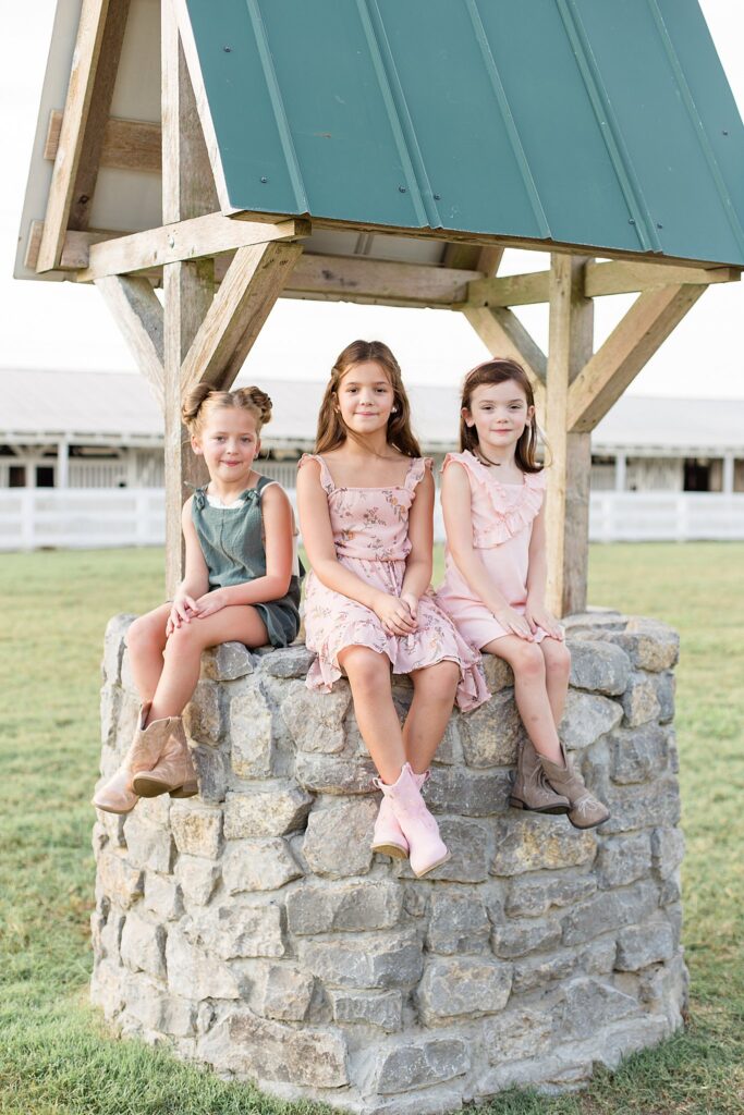 3 sisters sit on a rock well at Harlinsdale Farm in Franklin, TN wearing coordinating outfits with cowgirl boots and light pink and teal colors.