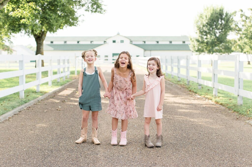 Wisp + Willow Photography Co, capture 3 sisters during their sibling photography session in Franklin, TN.  They hold hands and laugh at the camera as they all coordinate with their matching cowgirl boots and light pink and teal outfits!