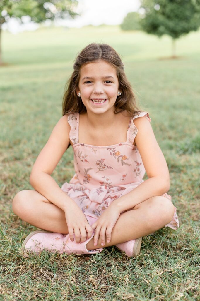Little girl sits criss cross applesauce on the grass as she wears a light pink dress with florals and smiles at the camera of Wisp + Willow Photography during their sibling photography session ini Franklin, TN.
