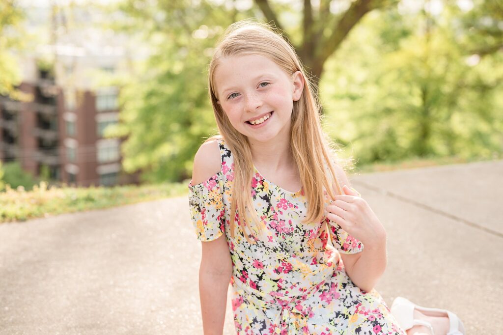 Young girl wears a floral, open shoulder, spring dress as she sits on the sidewalk and holds her hair with one hand as she smiles at the camera during her family photography session in Richmond, VA.