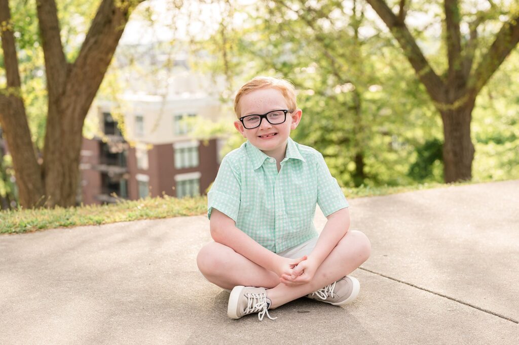 Young boy wears glasses and sits criss cross applesauce with his mint and white button shirt and khaki shorts/
