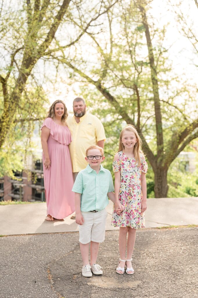 Daughter and son stand in the forefront of the image as they hold hands and smile at the camera of Wisp + Willow Photography Co., while Mom and Dad stand in the back holding hands and looking at their kids.