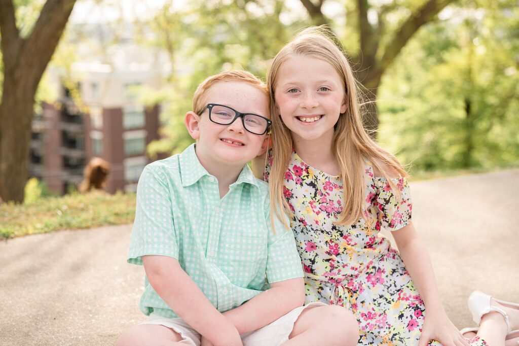 Brother and sister sit next to each other on the sidewalk at Libby Hill Park during their family session in Richmond.  They smile beautifully at the camera wearing spring colors during the beautiful springtime in Richmond.
