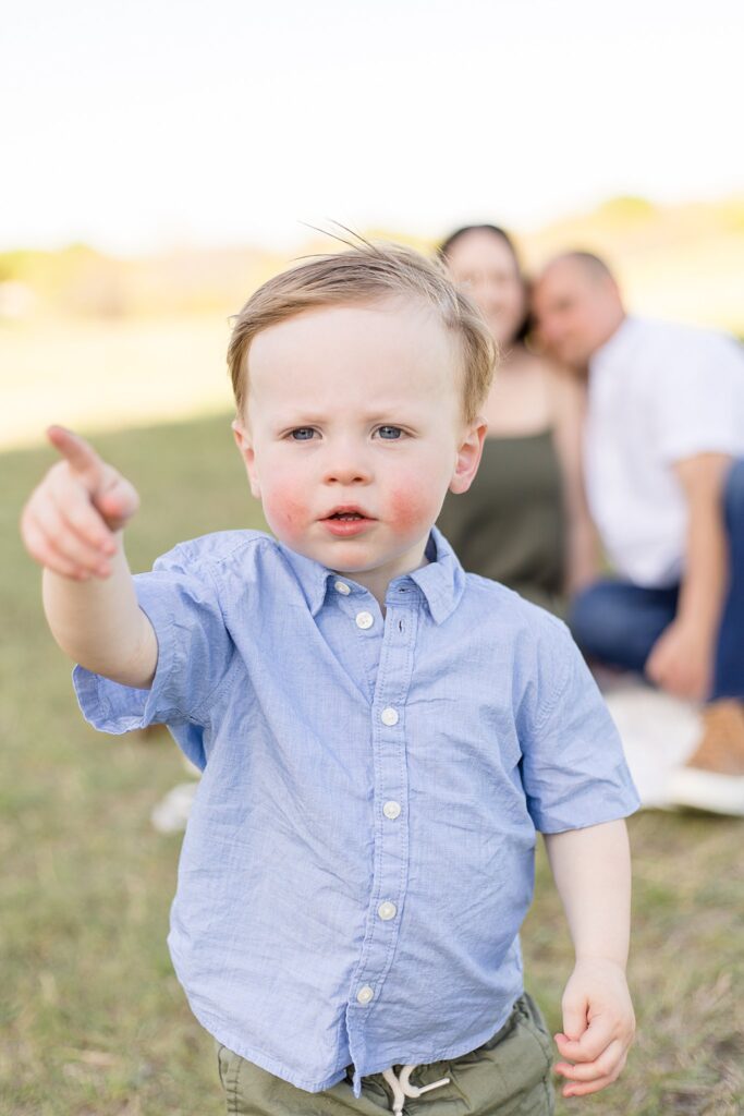 Toddler boy with rosy cheeks points off into the distance as mom and dad are in the background blurred out smiling at their son during their family photography session with Wisp + Willow Photography Co.