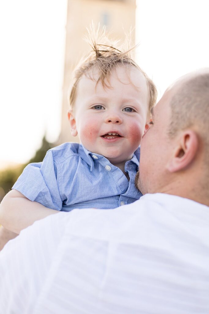 Rosy cheek toddler boy smiles over his Dad's shoulder at family photographer, Wisp + Willow Photography Co. 's camera during their family portrait session.
