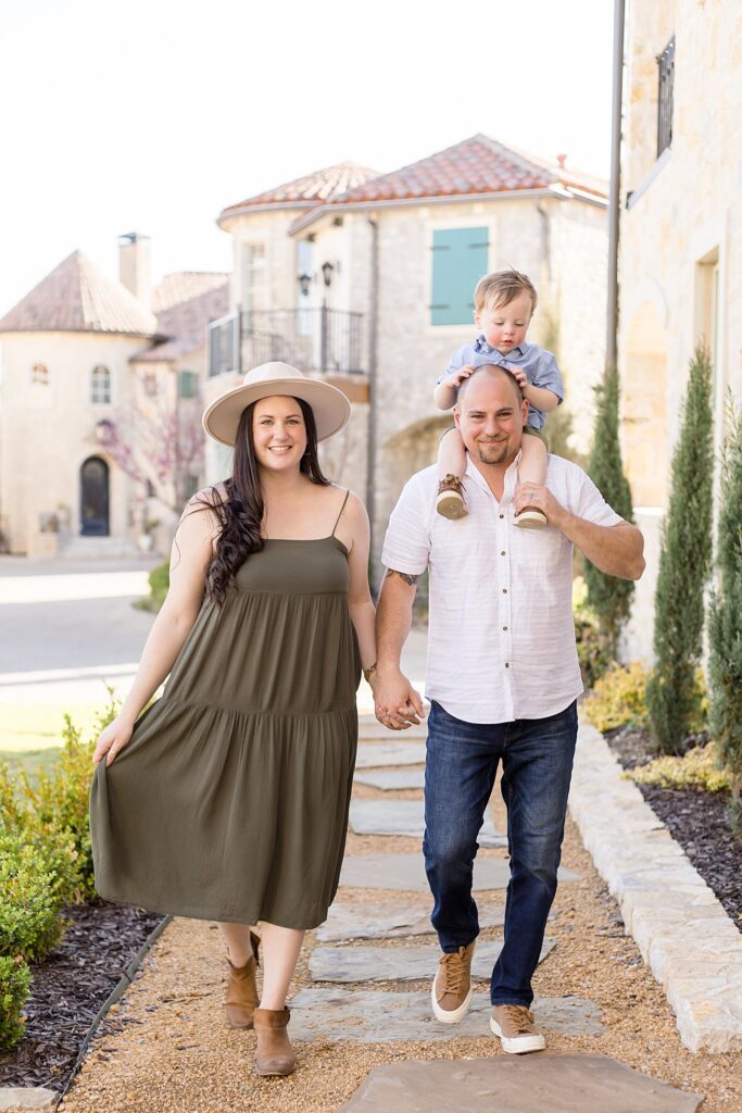 Dad puts son on his shoulders as he walks holding hands with his wife as they smile at the camera walking down the cobblestone pathway at Adriatic Village.