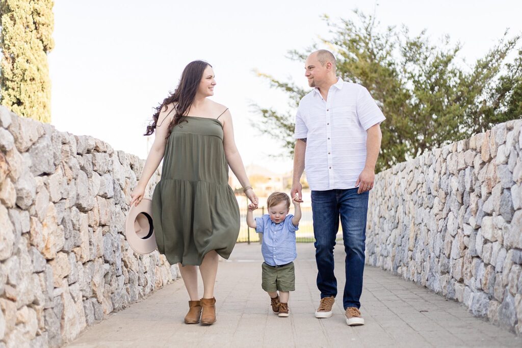 Mom and Dad hold toddler son's hands as they walk down a stone wall pathway looking at each other during their McKinney family photography session.