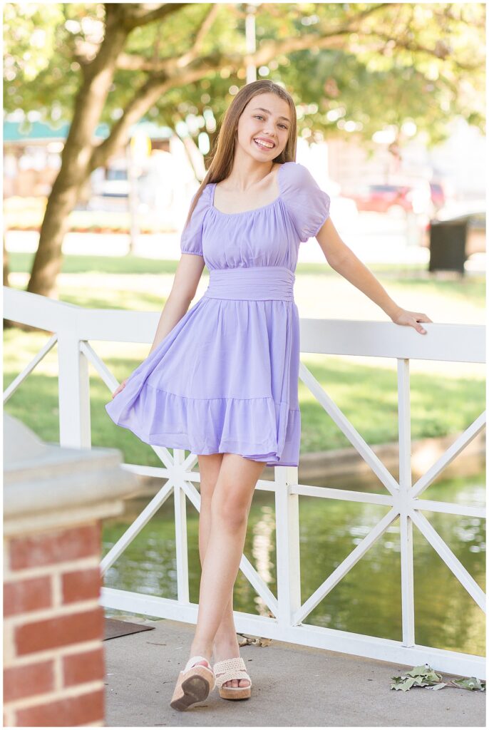 Senior session in Plano, TX of girl with blonde hair and wears a purple dress, cream lace wedges and smiles at camera of Wisp + Willow Photography Co.