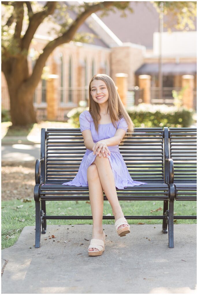 Senior session in Plano, TX has girl sit on black slated metal bench as she wears a purple dress and has her hands on her knees.