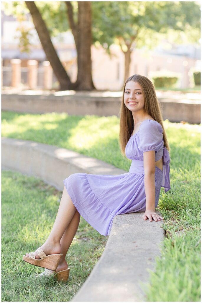 Plano senior photographer, Wisp + Willow Photography Co. capture senior girl who wears a purple chiffon dress as she sits on concrete wall surrounded by grass and smiles at the camera.
