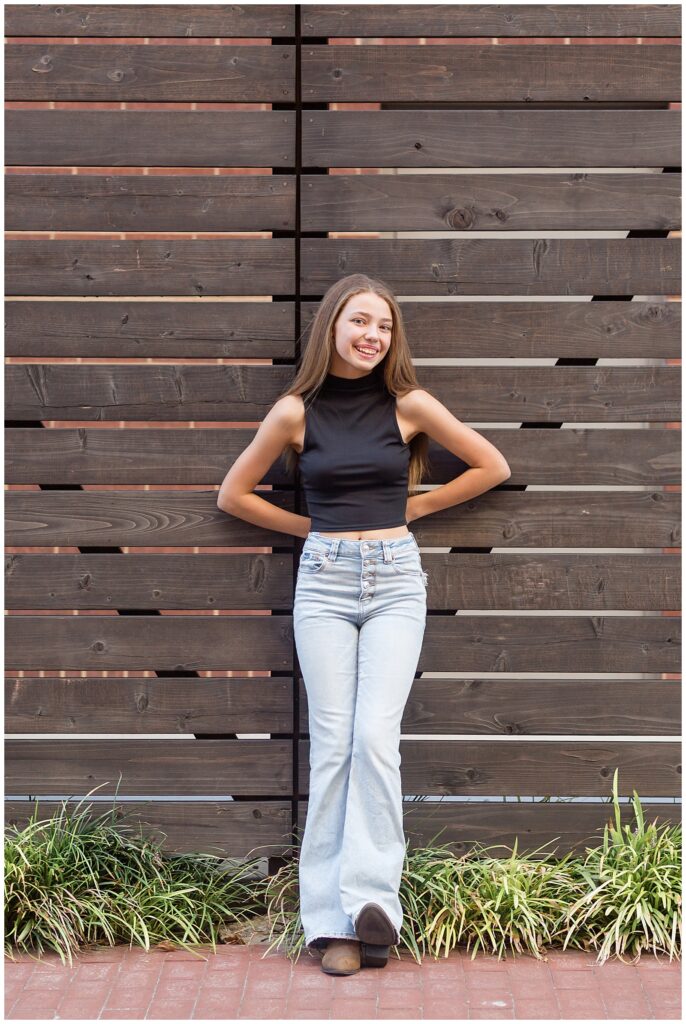 Senior portrait session in Downtown Plano, TX is of a senior girl who wears a black, sleeveless, crop top and bell bottom jeans with cowgirl boots.