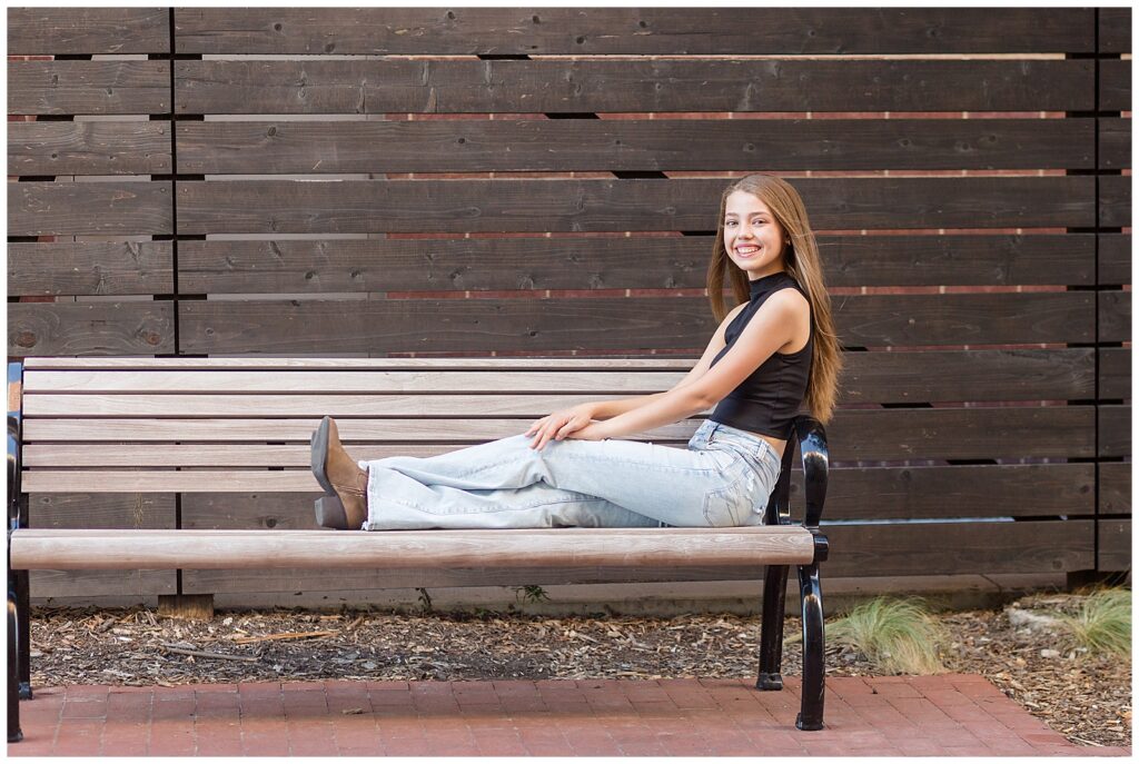 Senior sits on bench with legs up and her hands on her knees as she smiles at the camera for her senior portrait session in Downtown, Plano, TX.