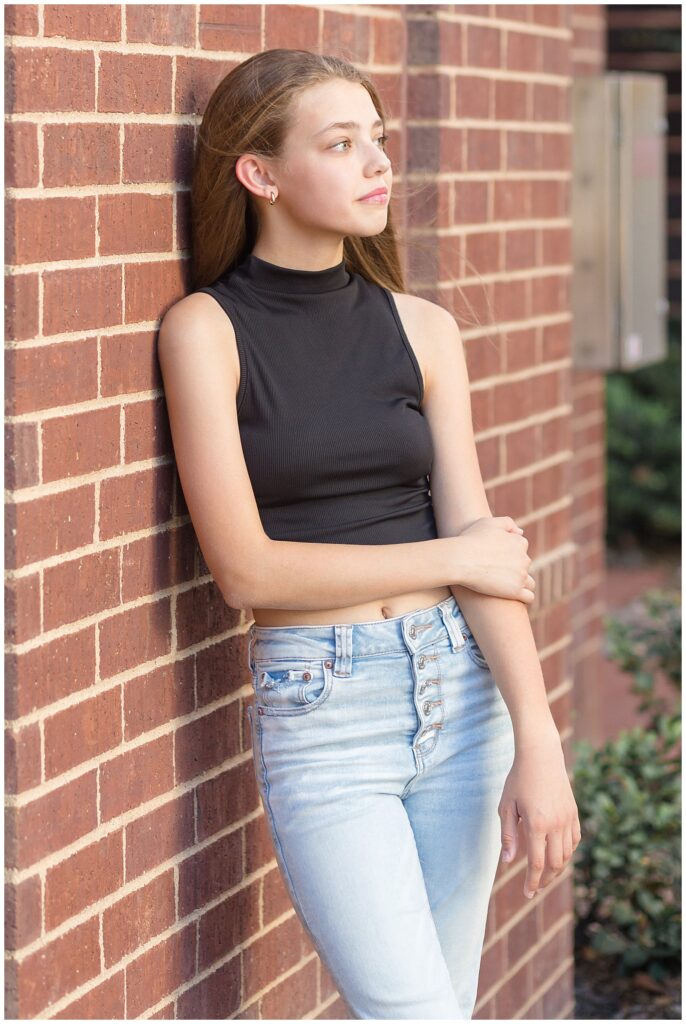 Girl leans against brick wall as she looks off into the distance wearing a black crop top with a mock neckline and no sleeves during her senior portrait session.