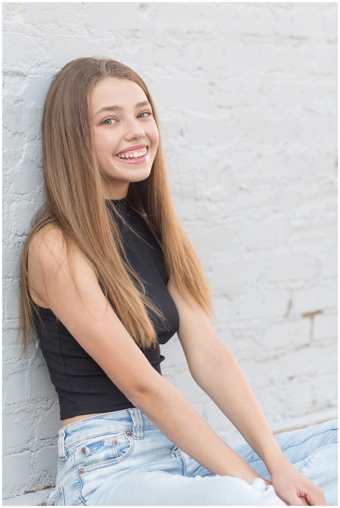 Beautiful image of senior girl from Plano, TX smiles at the camera of Wisp + Willow Photography Co. as she sits against a white brick wall.