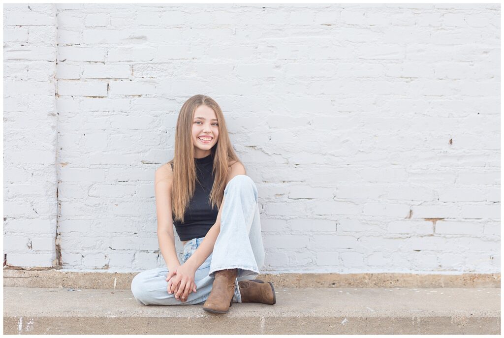 Girl sits with one leg under her other as she sits against a white brick wall and leaves her hair down for her senior portrait session in Plano, TX.
