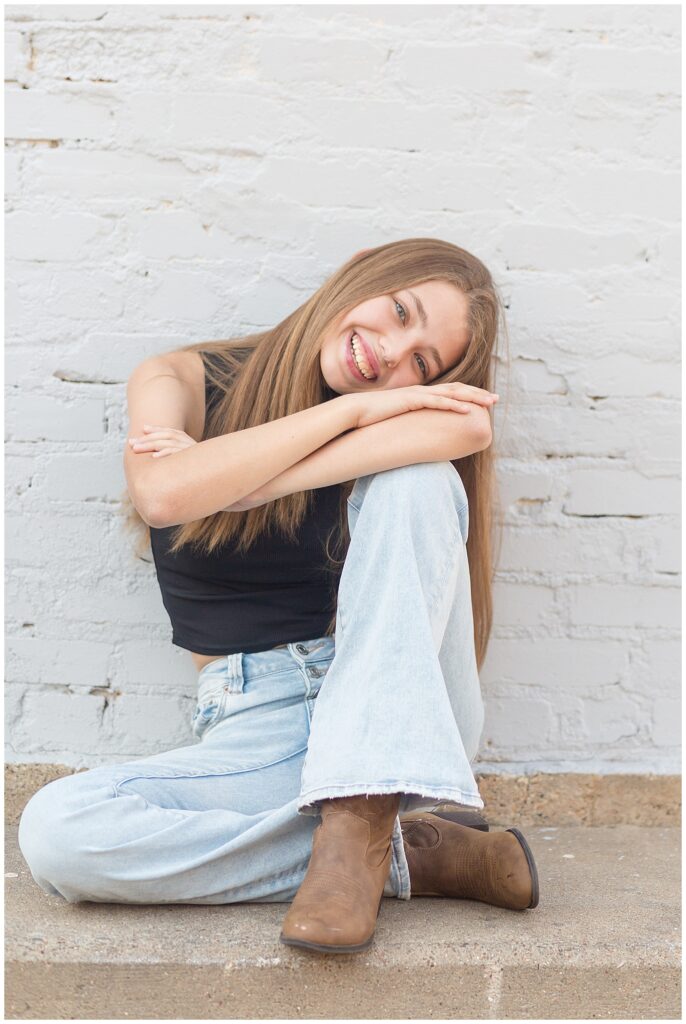 Senior girl puts one knee up and rests her head on her arms as she sits against a white brick wall and smiles at the camera of Wisp + Willow Photography Co.