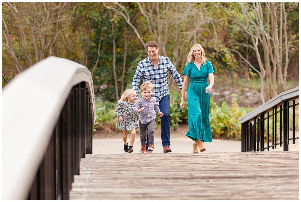 Family of 4 walk across bridge as they have some fun and laugh together as the kids run ahead.