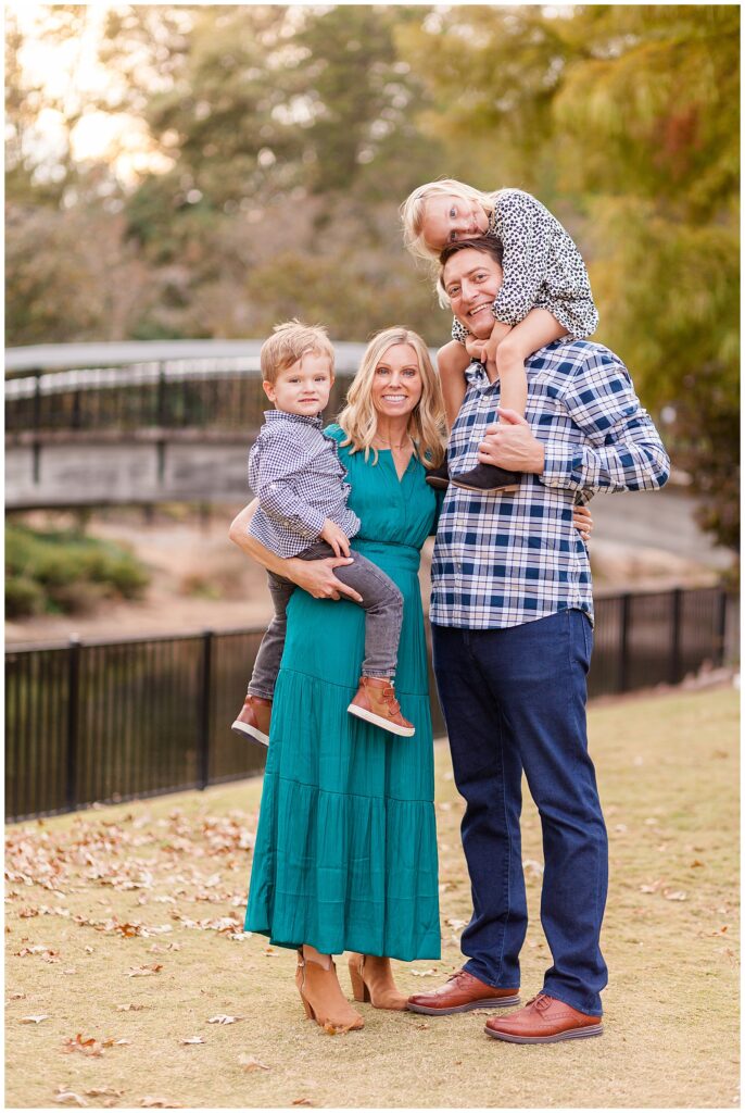 Raleigh family photographer, Wisp + Willow Photography Co., take a family portrait of a family of 4 as daughter sits on Dads shoulders and rests her head on his head and Mom holds son on her hip as they smile at the camera.