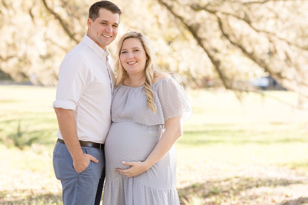 Savannah maternity session at Wormsloe Historic Site has soon-to-be dad wearing a white button down shirt, brown belt, and grey slacks while mom wears a flowly, eyelet grey dress as they both smile at the camera during their session.